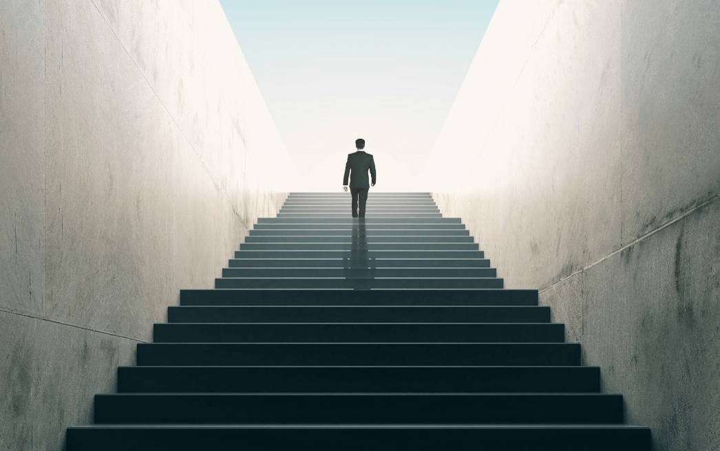 A stock photo of someone walking up a set of stairs into the distant sunlight.