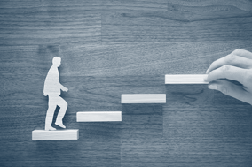 Stock photo of a hand placing four shelves arranged as stairs. On the first step is a mini-cut-out of a person walking.