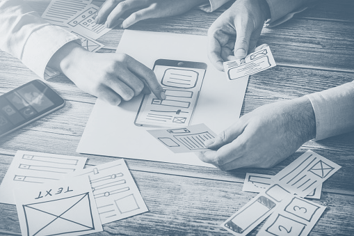 Stock photo of multiple hands placing app/website design elements on a piece of paper.